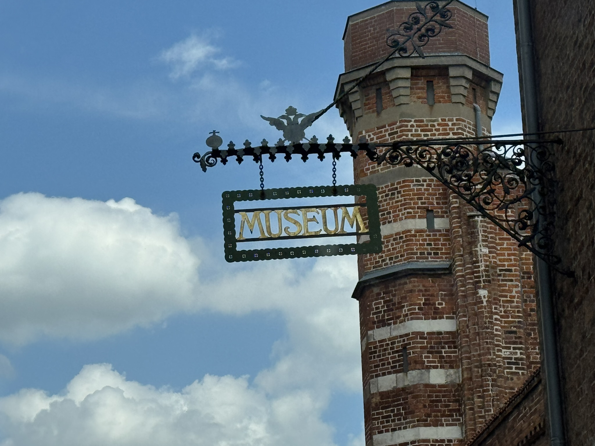 Ein hängendes Schild mit der Aufschrift "Museum". Dahinter ist ein Turm aus Backstein und der Himmel zu sehen.