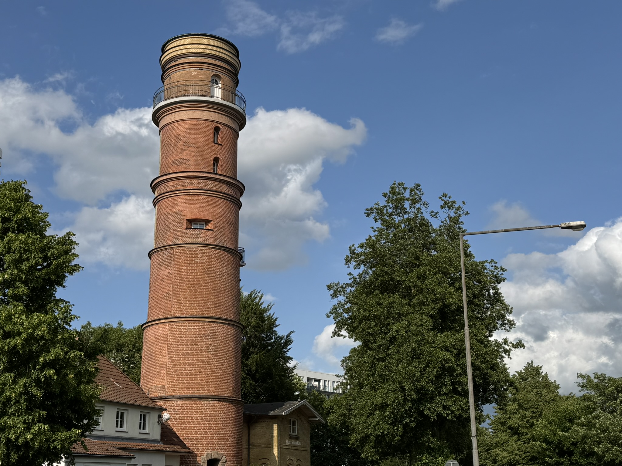 Ein Leuchtturm aus braunen Ziegeln in mitten einer grünen Landschaft mit einem Hochhaus im Hintergrund.
