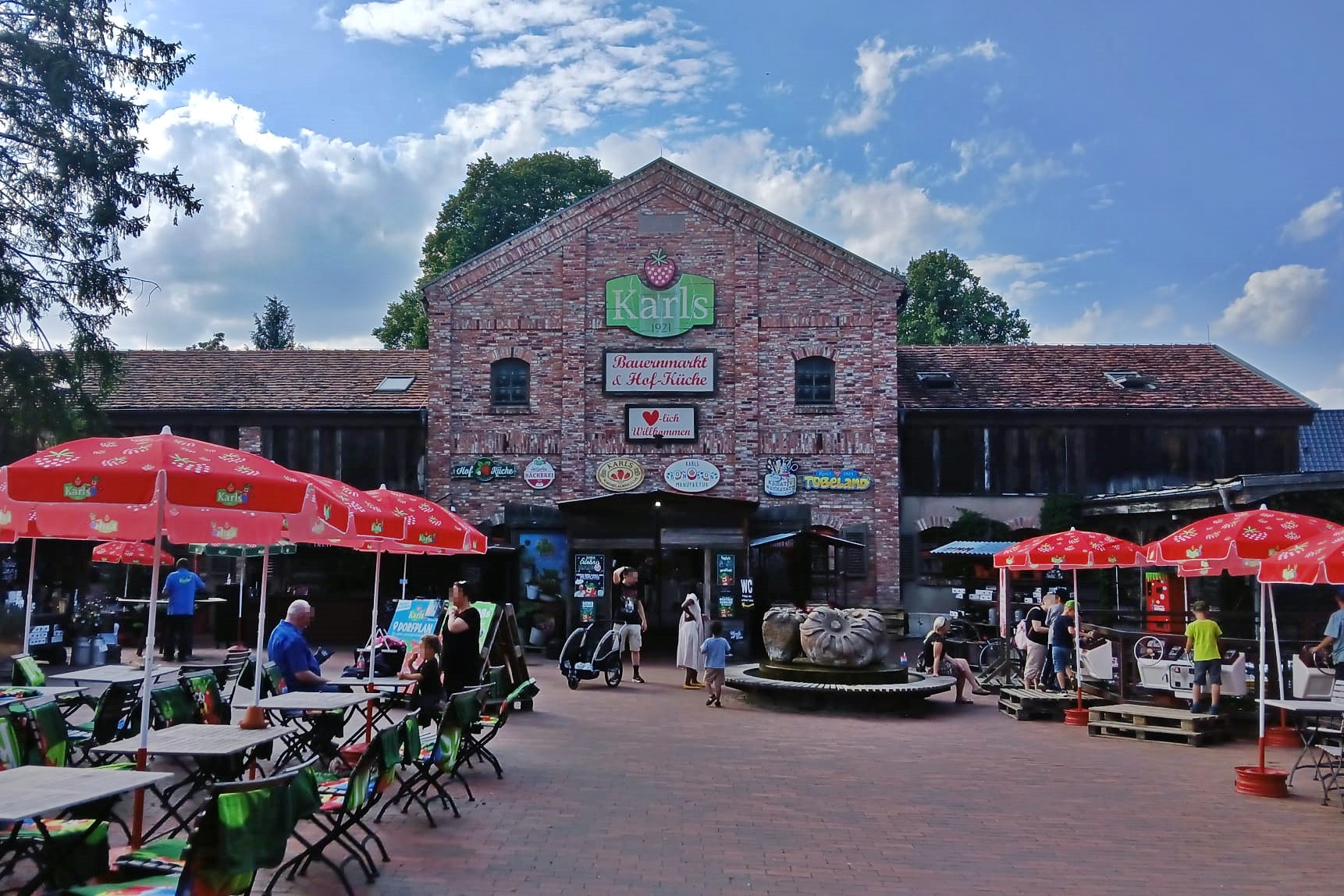 Der Vorplatz von Karls Erlebnis-Dorf in Warnsdorf. Ein Backsteingebäude mit vielen roten Sonnenschirmen mit Erdbeer-Muster drauf davor.